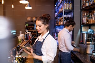Pretty young waitress with tablet scrolling through online orders while her colleague preparing tea for guests on background