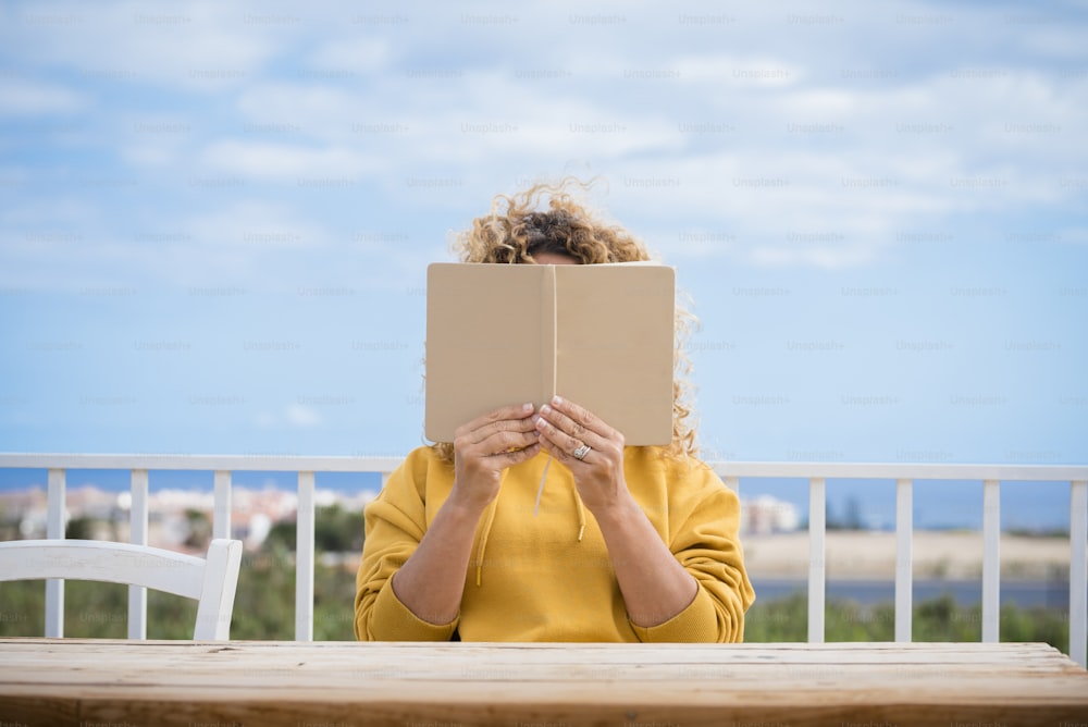 Femme méconnaissable lisant un livre en papier à l’extérieur avec une table en bois devant elle et un beau ciel bleu en arrière-plan - concept de lecture et d’étude pour les gens