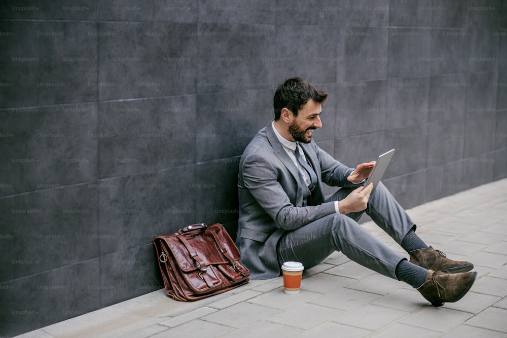 Jeune homme d’affaires attrayant à la mode assis dans la rue et utilisant une tablette.