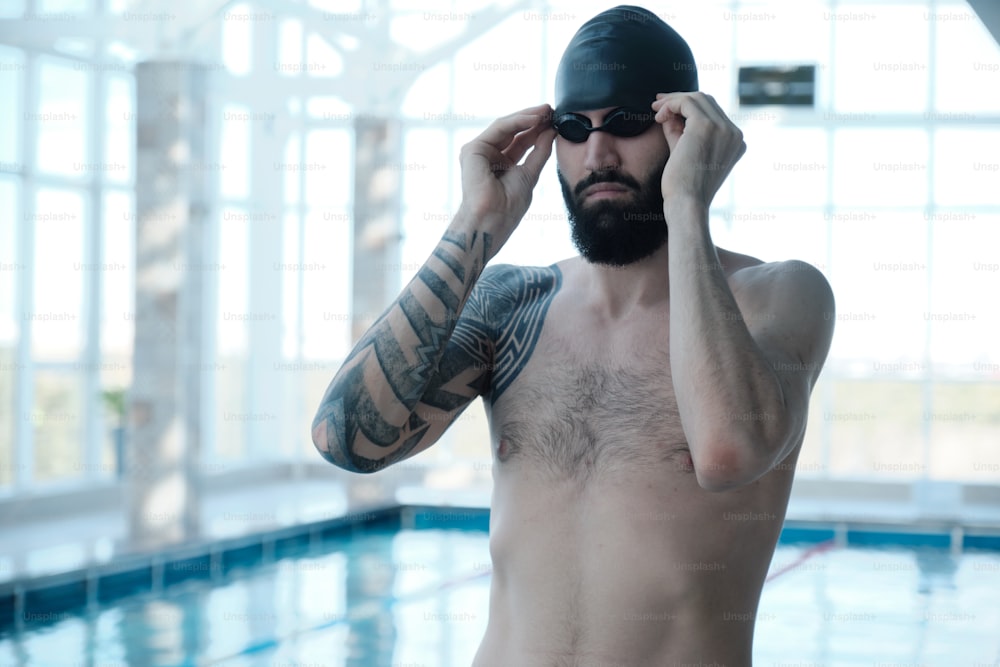 Brutal bearded swimmer with tattoo adjusting goggles while preparing for swimming training