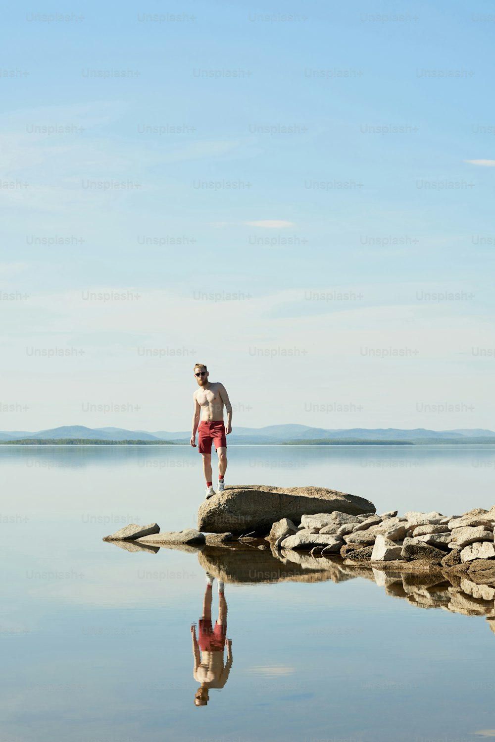 Young shirtless man in sunglasses standing on large stone against beautiful serene lake on bright summer day