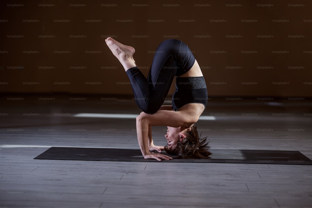 Vista lateral de la mujer yogui de ajuste flexible que se balancea sobre la cabeza. Interior del estudio de yoga.