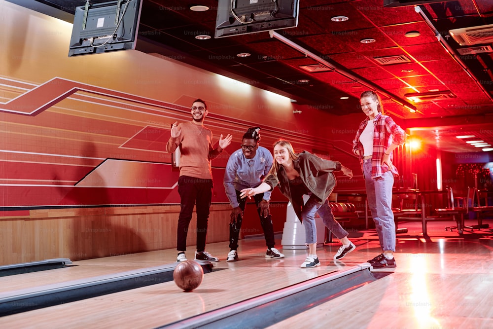 Group of young cheerful friends of various ethnicities standing by bowling alley while obne of them throwing ball during game