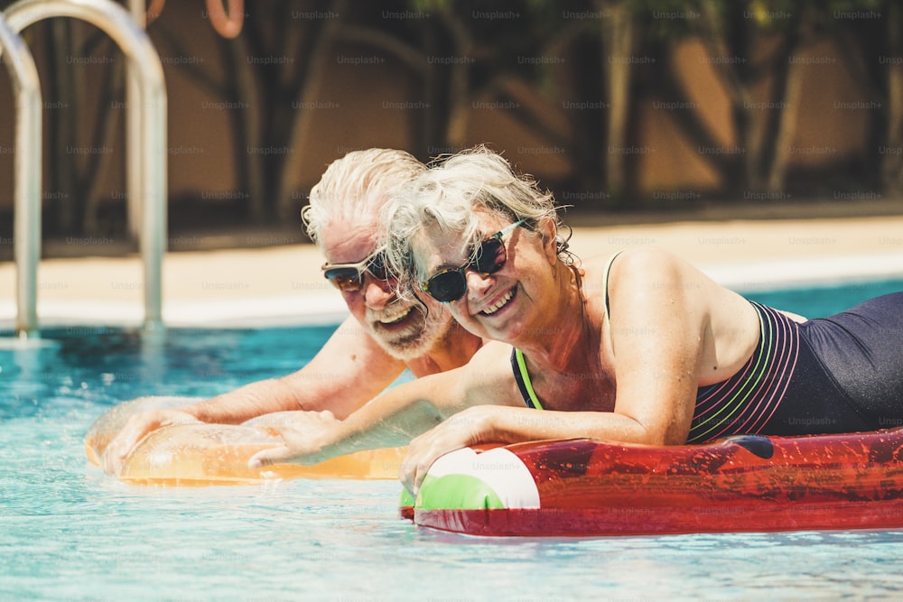 Cheerful happy coule of retired senior old people smile and enjoy together the pool in summer hoiday vacation with coloure trendy lilos in the water - concept of no limit age to have fun and love