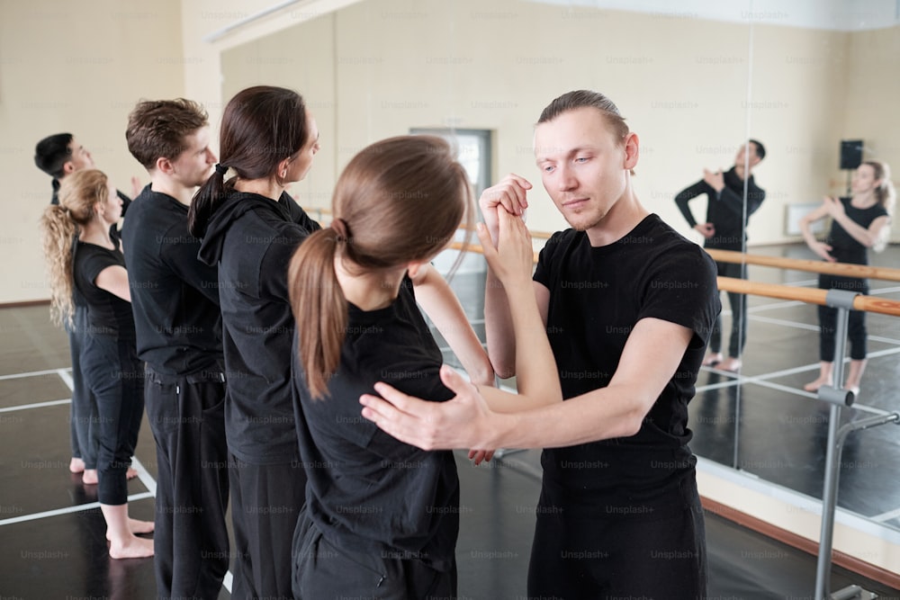 Young instructor of modern ballet dancing cource standing by one of his students while helping her with exercise at lesson
