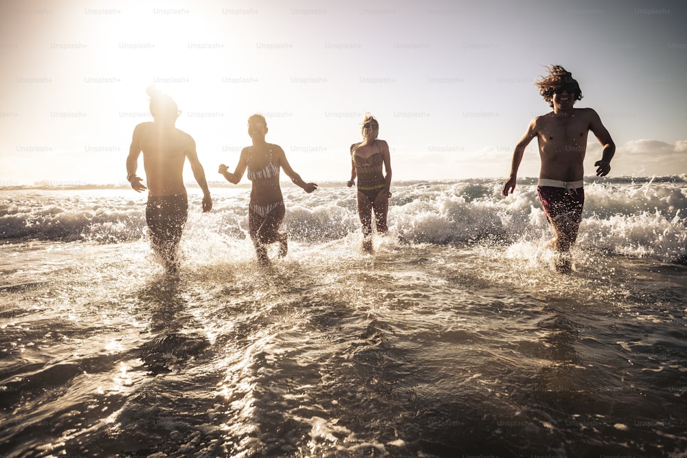 Jovens amigos correm na água do mar juntos se divertindo em um dia ensolarado de férias de verão - pessoas em atividade de lazer ao ar livre brincando com ondas e oceano