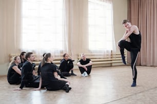Group of several young dance course students sitting on the floor while looking at guy in activewear doing exercise