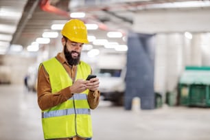 Jovem trabalhador da construção civil barbudo sorridente em pé na garagem subterrânea em processo de construção e usando o telefone inteligente para mensagens de texto. É hora do intervalo.