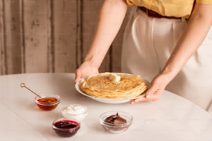 Hands of young housewife putting plate with stack of hot homemade pancakes on table with honey, sourcream, jam and chocolate