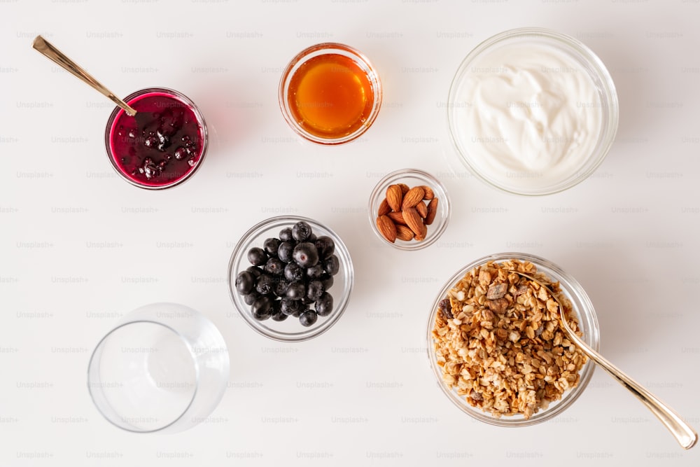 Flat layout of white kitchen table with several bowls containing muesli, cherry jam, sourcream, almond kernels, honey and fresh blackberries