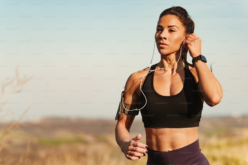 Young athletic woman adjusting earphones while exercising and running in nature. Copy space.