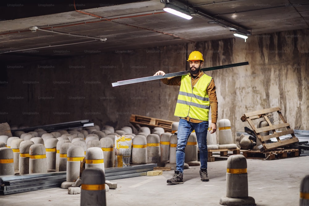 Full length of handsome bearded worker in working clothes carrying metal profile while walking in underground garage in construction process.