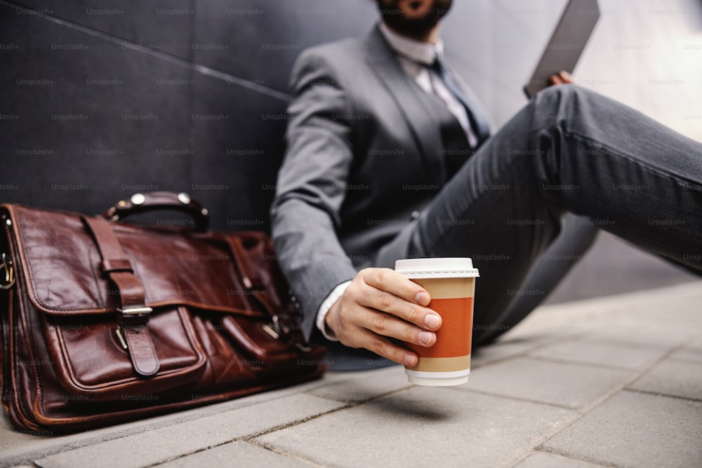 Nahaufnahme eines jungen Geschäftsmannes, der auf der Straße sitzt, ein Tablet benutzt und eine Einwegtasse mit Kaffee hält. Selektiver Fokus auf die Hand.