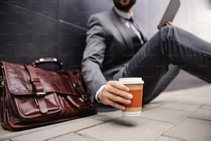 Primer plano de un joven hombre de negocios sentado en la calle, usando una tableta y sosteniendo una taza desechable con café. Enfoque selectivo a mano.
