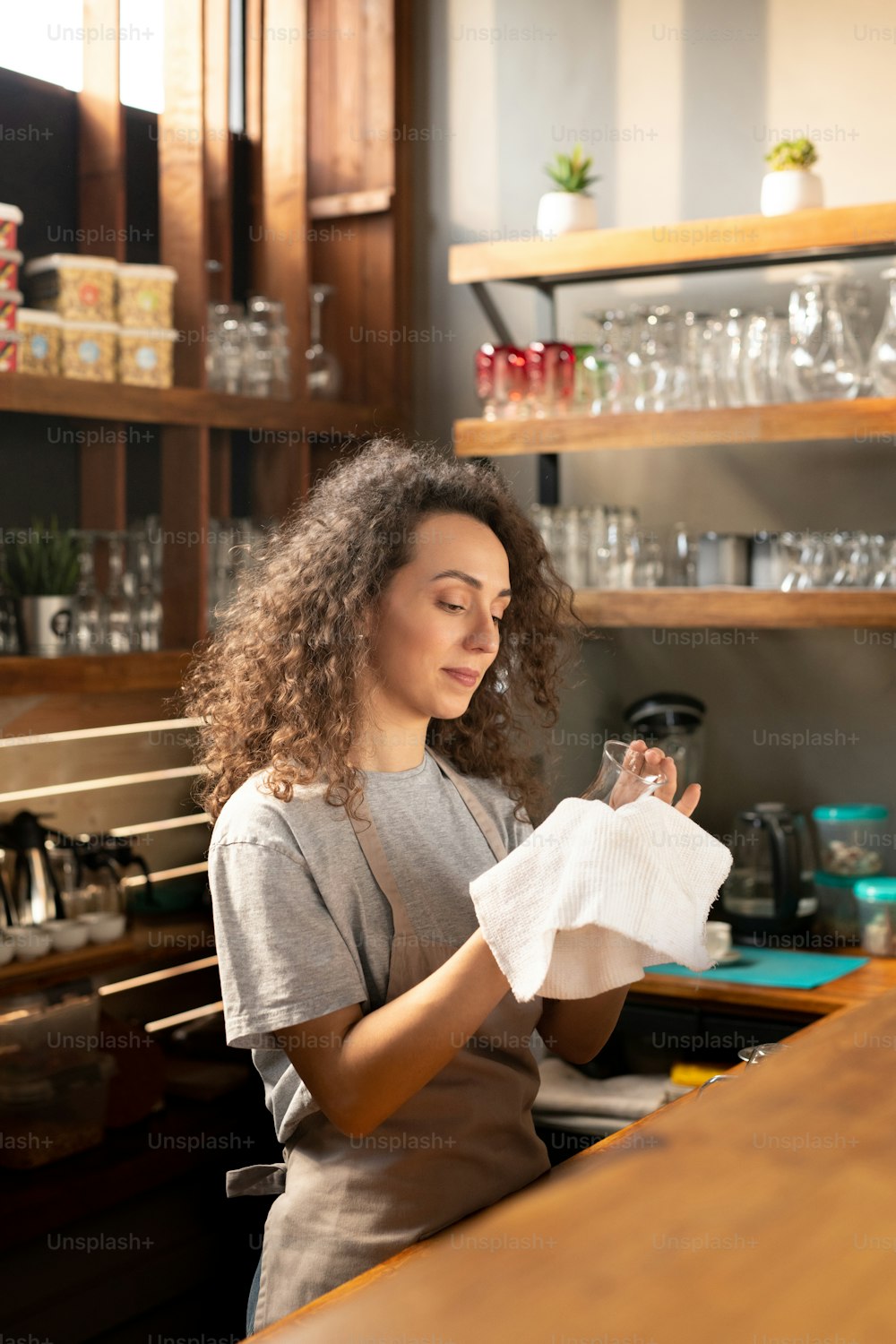 Pretty young waitress in workwear cleaning glass with white cotton napkin or towel while standing by bar counter in restaurant or cafe