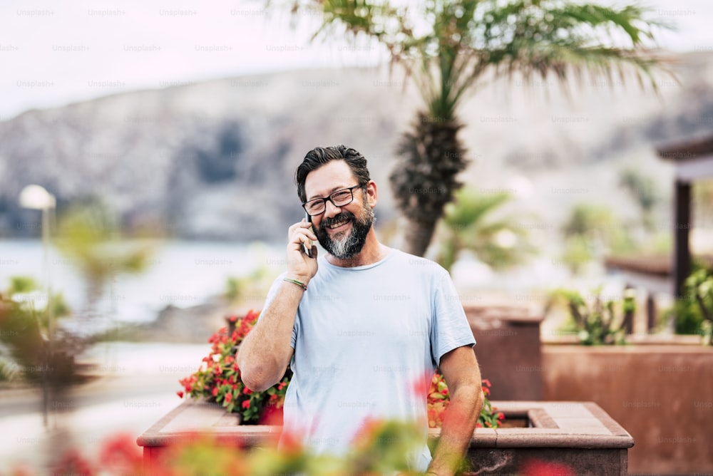 Cheerful adult senior man smile and do a phone call looking at the camera - outdoor defocused natural background - people and technology concept