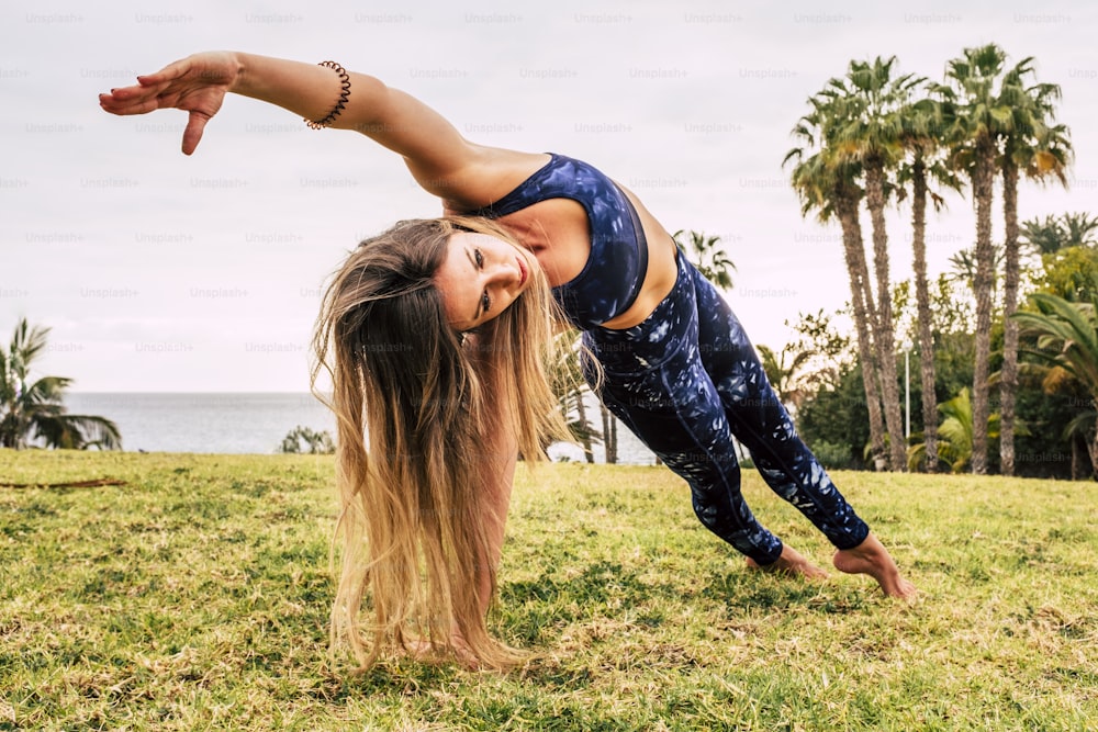 Active lifestyle people concept with beautiful long blonde hair young woman doing balanced pilates position on the green natural meadow in outdoor sport healthy activity - young female and fitness outdoors