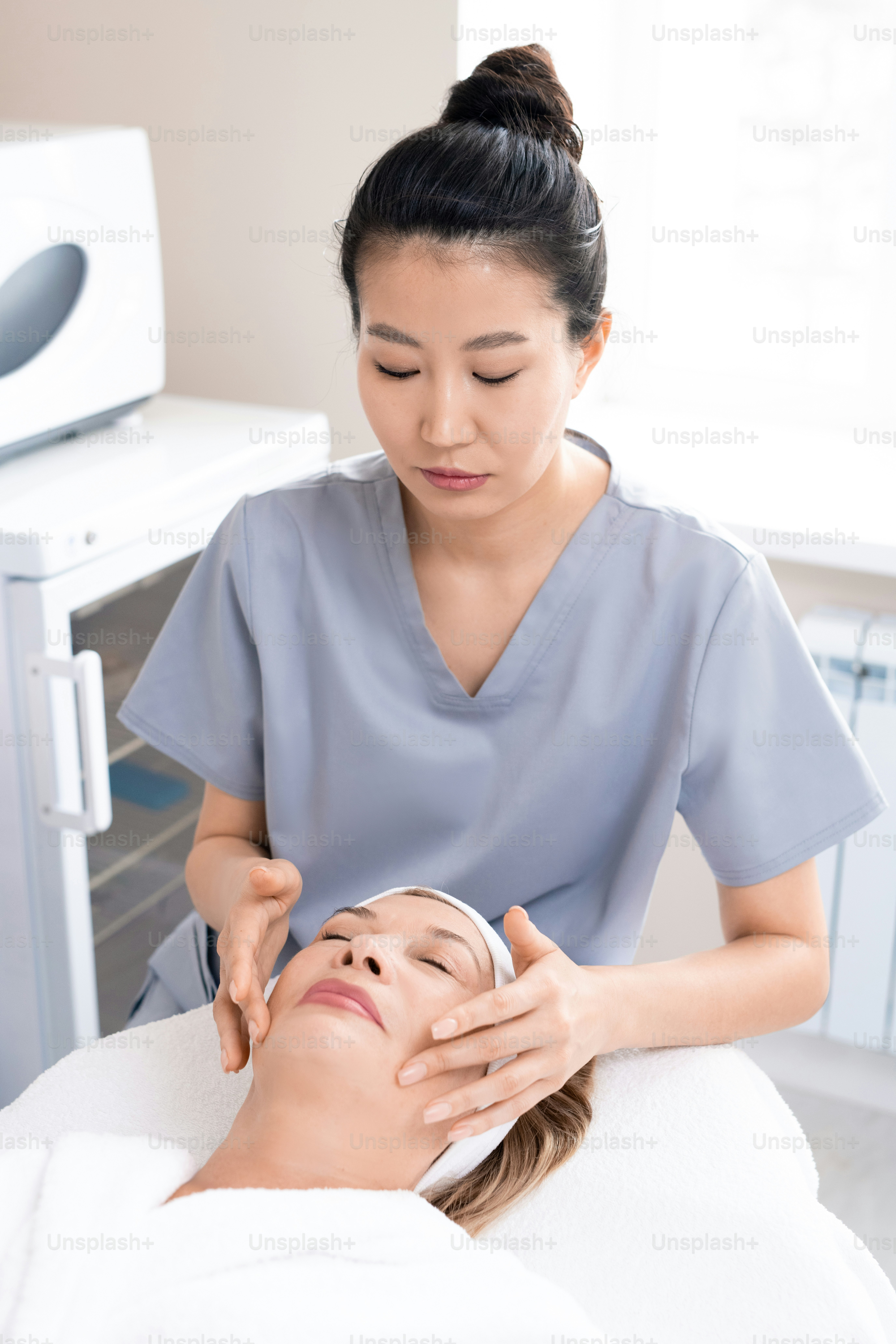 Masajista asiático concentrado que esculpe la cara de una mujer madura para reducir las arrugas en el procedimiento de tratamiento de belleza