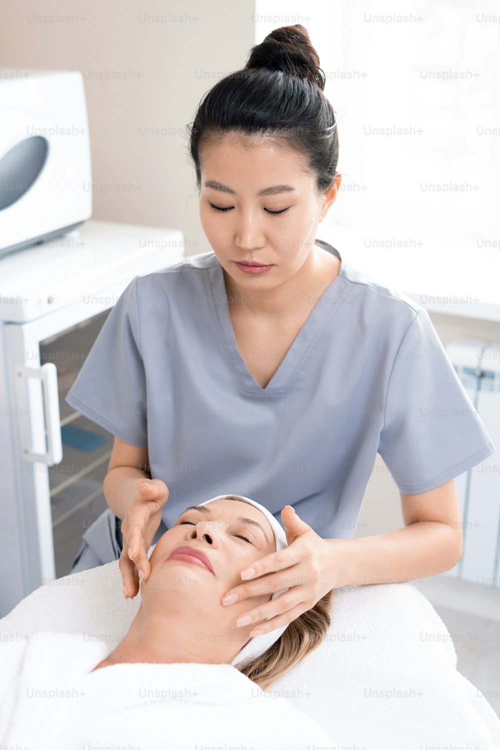 Concentrated Asian masseur sculpting face of mature woman to reduce wrinkles at beauty treatment procedure