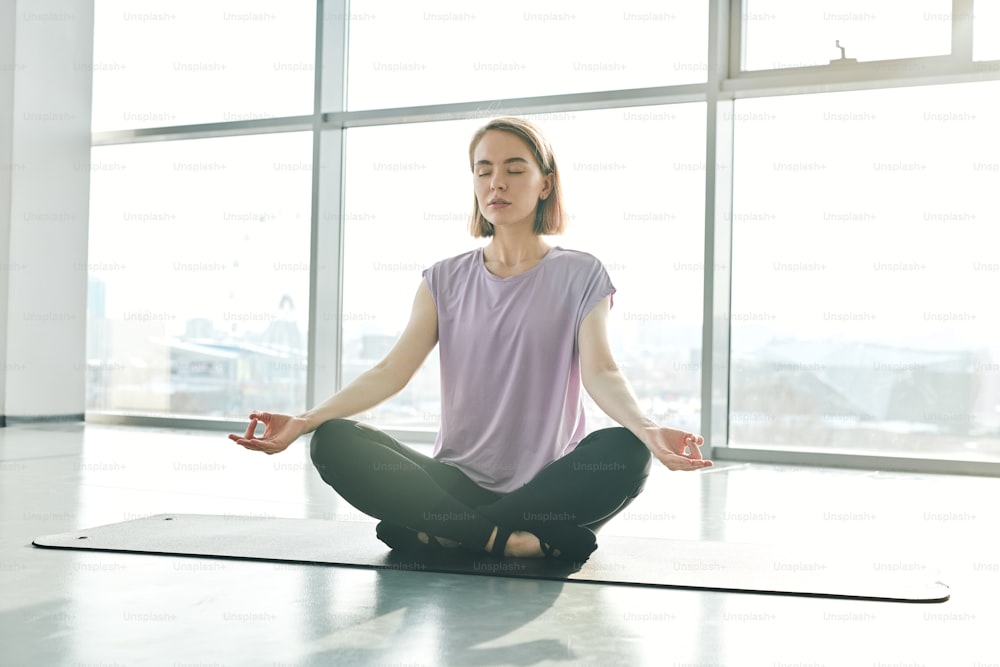 Pretty young serene woman in activewear sitting on mat while practicing lotus position during yoga training in gym or leisure center