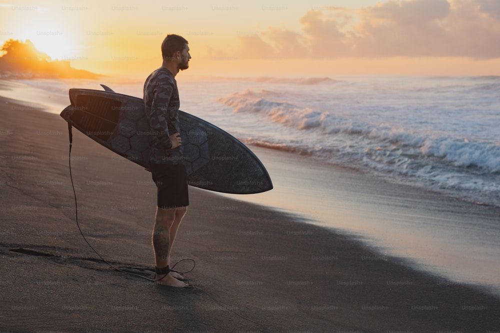 Surfing. Surfer With Surfboard Standing On Ocean Beach. Man Silhouette At Beautiful Sunrise In Bali.