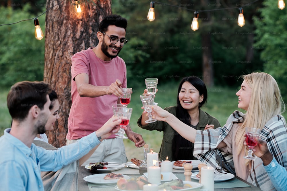 Three happy young intercultural couples with wineglasses sitting by served table and toasting for friendship during outdoor dinner