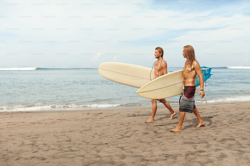 Surf. Surfistas guapos con tablas de surf. Hombres jóvenes caminando en la playa de arena. Estilo de vida activo, deporte acuático en un hermoso fondo oceánico.