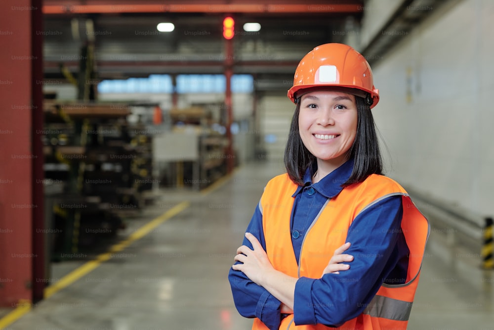 Retrato de un joven ingeniero asiático de fábrica confiado sonriente en casco naranja y chaleco de pie en el taller