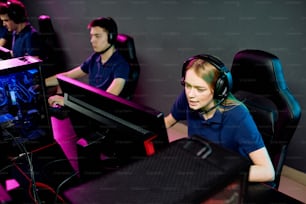 Bella ragazza dai capelli lunghi in auricolare che parla con qualcuno mentre è seduta davanti al monitor del computer e guarda lo schermo durante il videogioco