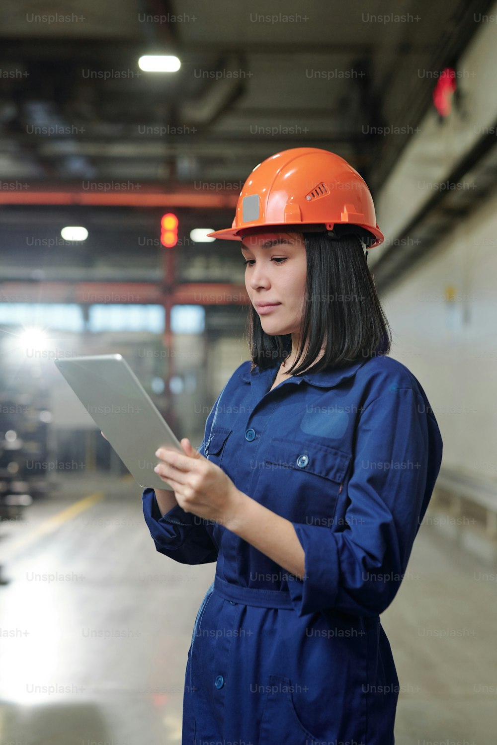 Serious young Asian female engineer in overall analyzing industrial processes with tablet app at factory