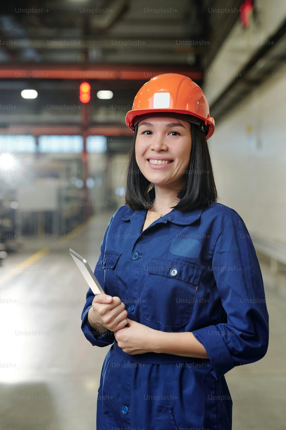 Portrait of positive young Asian woman in blue workwear and hardhat holding tablet in industrial shop