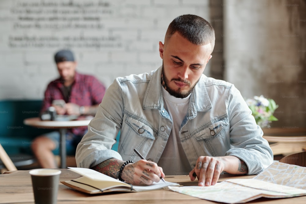 Konzentrierter Hipster-Typ mit Bart sitzt am Tisch in einem modernen Café und schaut sich eine Papierkarte an, während er sich Notizen über die Reise macht