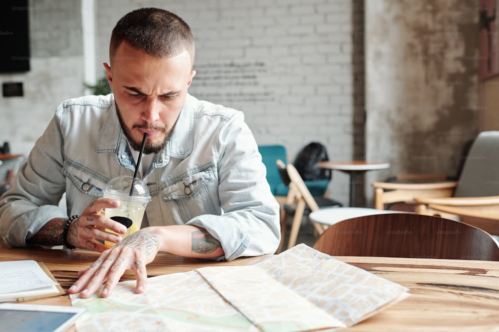 Frowning young bearded man in denim jacket sitting in cafe and drinking cold coffee while finding best places to visit using map