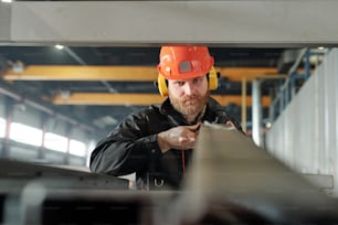 Serious bearded manual worker in ear protectors and hardhat sorting metal planks on shelf in warehouse