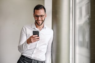 Young handsome smiling elegant man standing indoors and tying or reading message on smart phone.