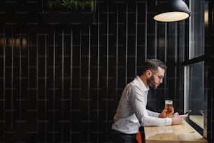 Vista lateral de un atractivo hombre barbudo de pie en el bar después del trabajo, tomando un vaso de cerveza y usando un teléfono inteligente para pasar el rato en las redes sociales.