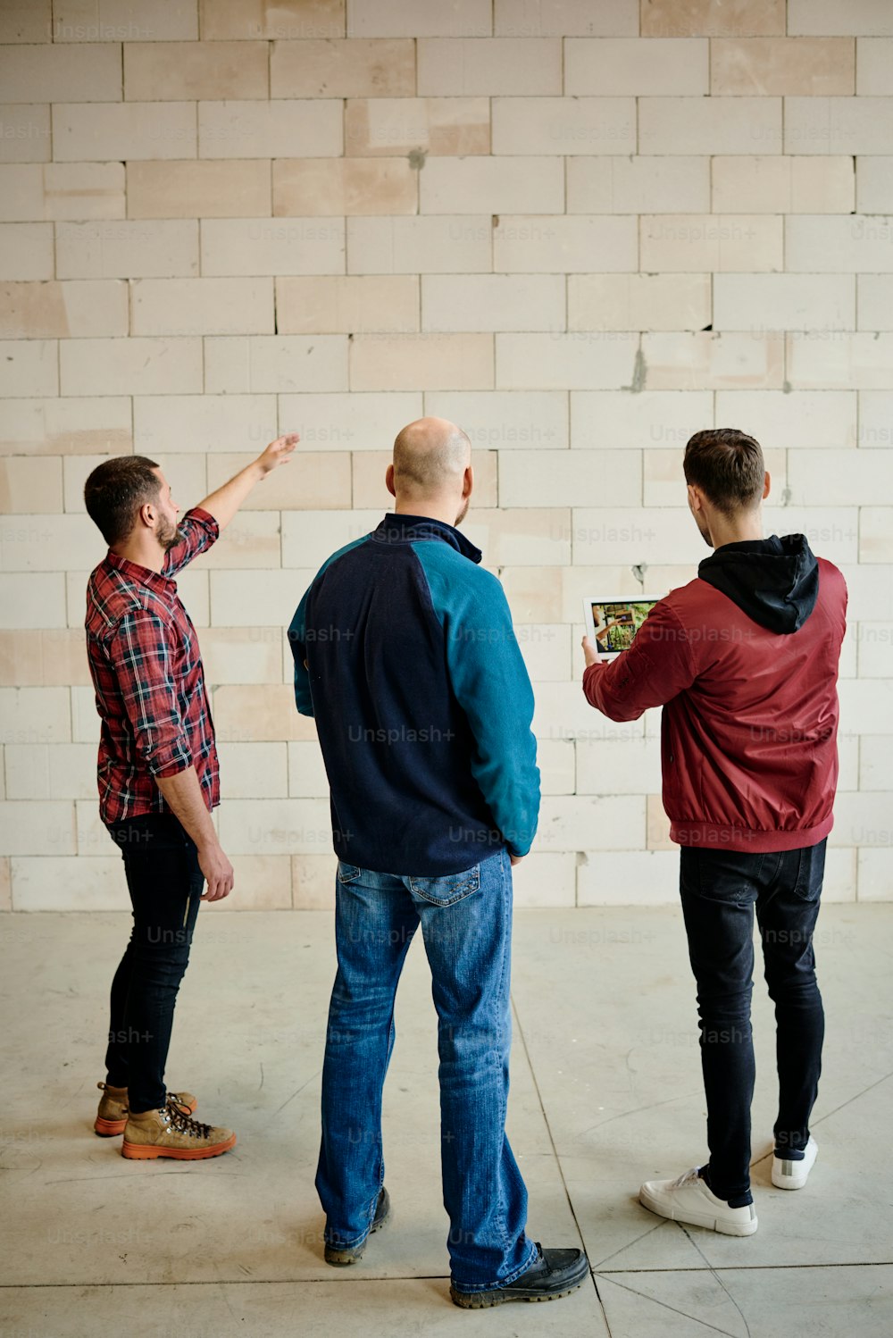 Vista posterior de tres jóvenes constructores o arquitectos discutiendo la calidad de los ladrillos que componen la pared de un edificio inacabado durante la reunión de inicio