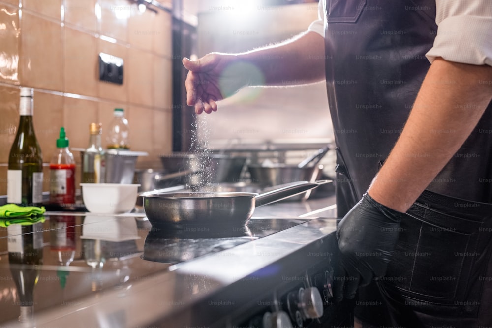 Close-up of unrecognizable chef in apron standing at stove and salting food in pan