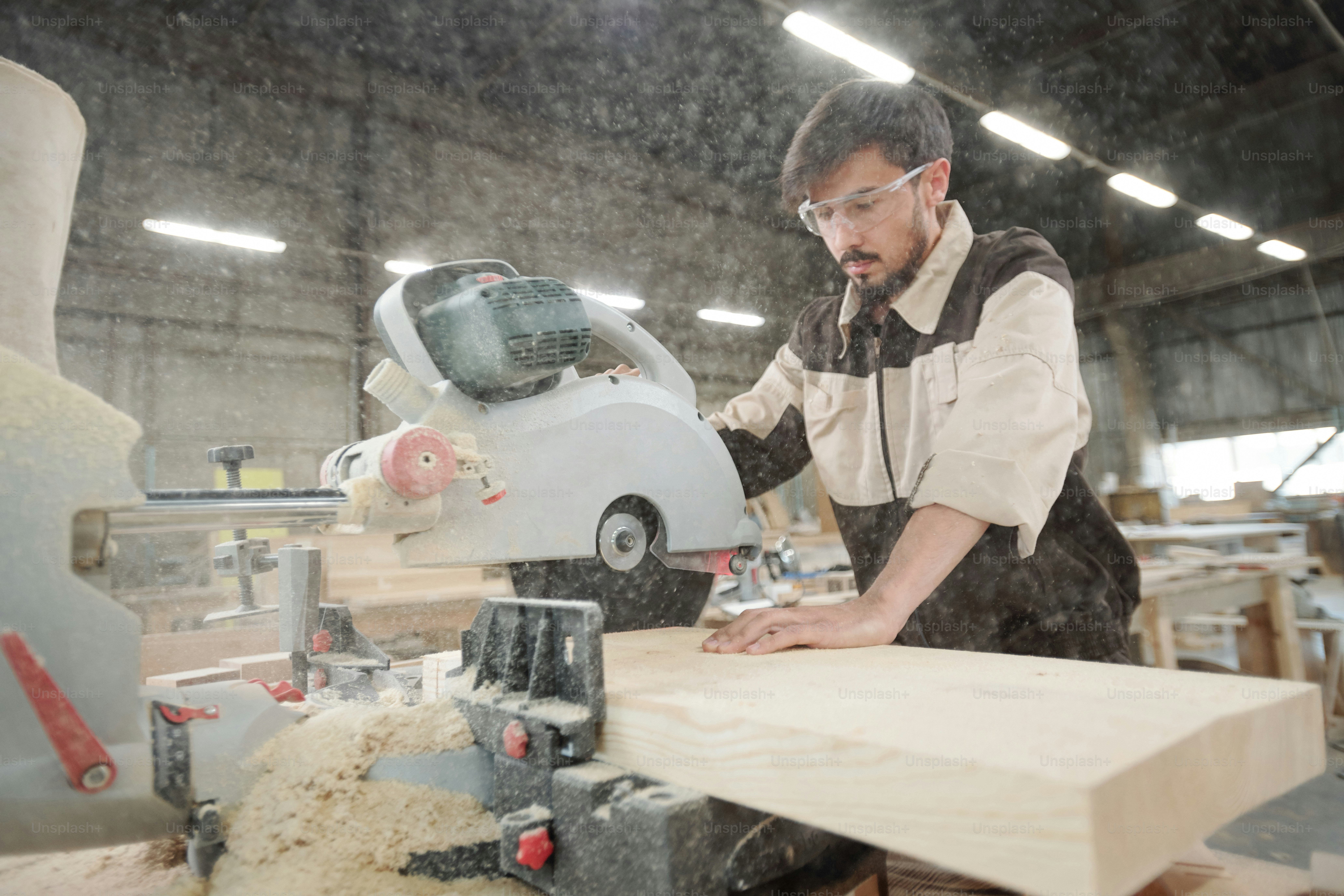 Young worker of furniture production factory in uniform and protective eyeglasses bending over large wooden board while using electric saw