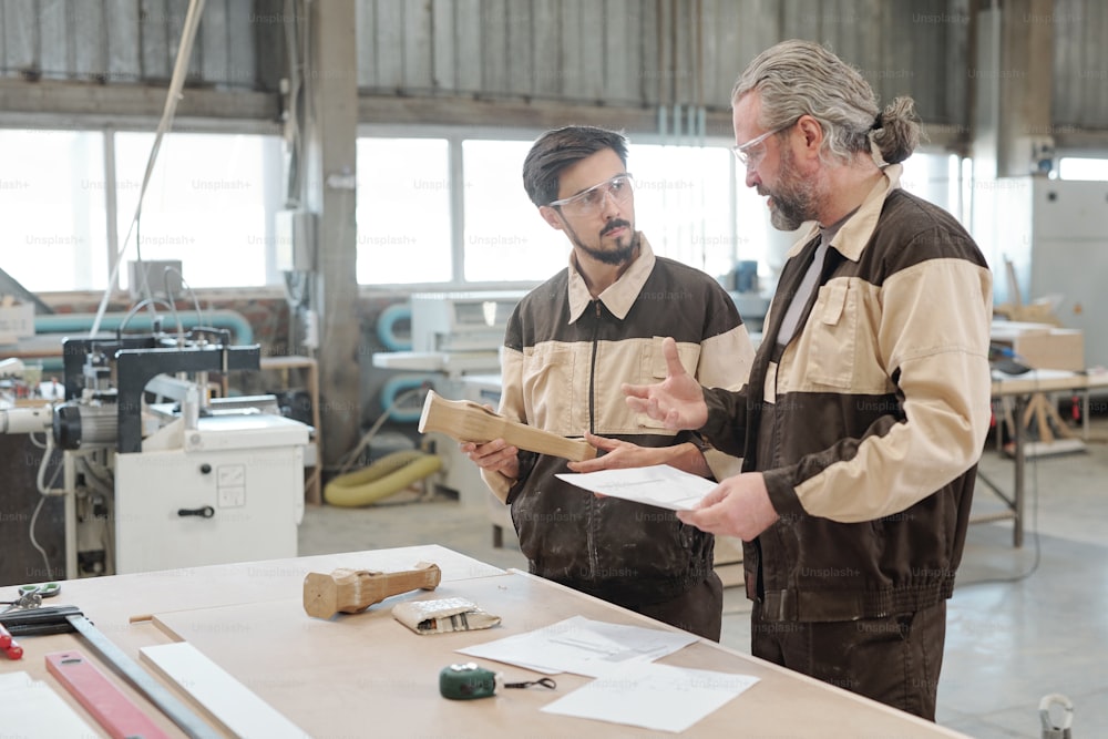 工場の作業場の大きなテーブルで、若い研修生に木製のワークピースのスケッチを説明する年配の専門技術者