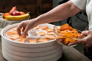 Hände der jungen Hausfrau, die Scheiben gelber getrockneter Früchte in eine Glasschüssel geben, während sie sie aus dem runden oberen Tablett des Lebensmitteltrockners nehmen