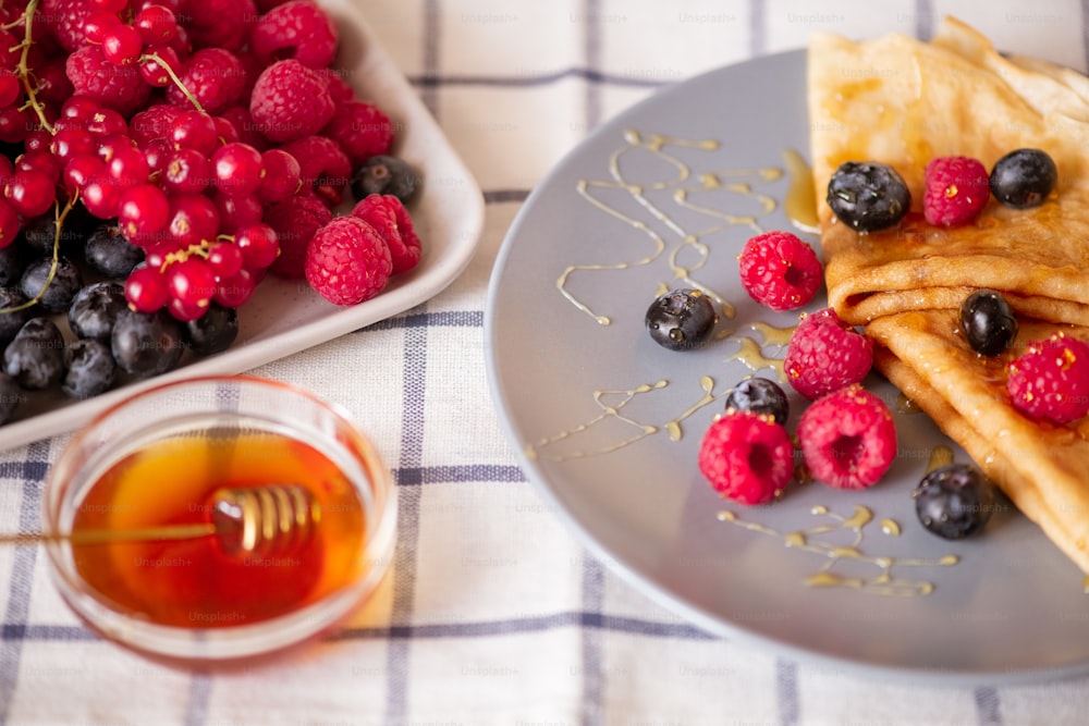 Two folded homemade pancakes with honey and fresh ripe raspberries and blackberries on grey porcelain plate on kitchen table