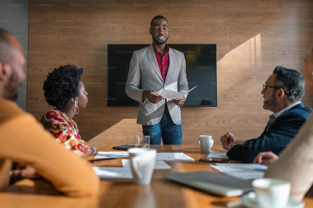Un jeune manager africain passe en revue la paperasse avec son personnel diversifié lors d’une réunion autour d’une table dans une salle de conférence de bureau