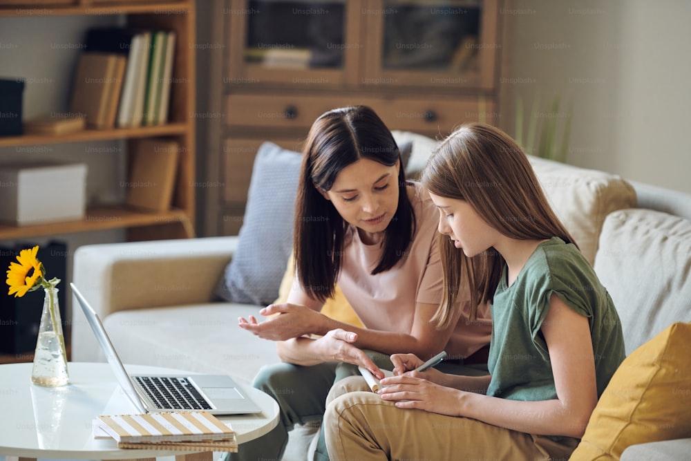 Middle-aged mother sitting on sofa and pointing at notes while helping daughter with homework