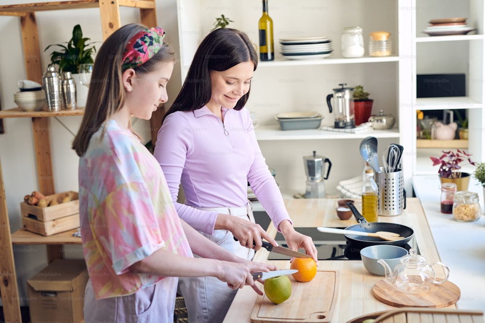 Positive middle-aged mother and her teenage daughter standing at kitchen counter and cutting fruits while making breakfast together