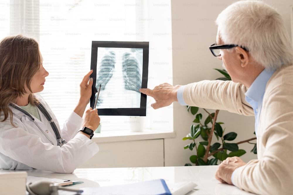 Senior patient and young female pulmonologist pointing at x-ray image of lungs while discussing their characteristics during consultation