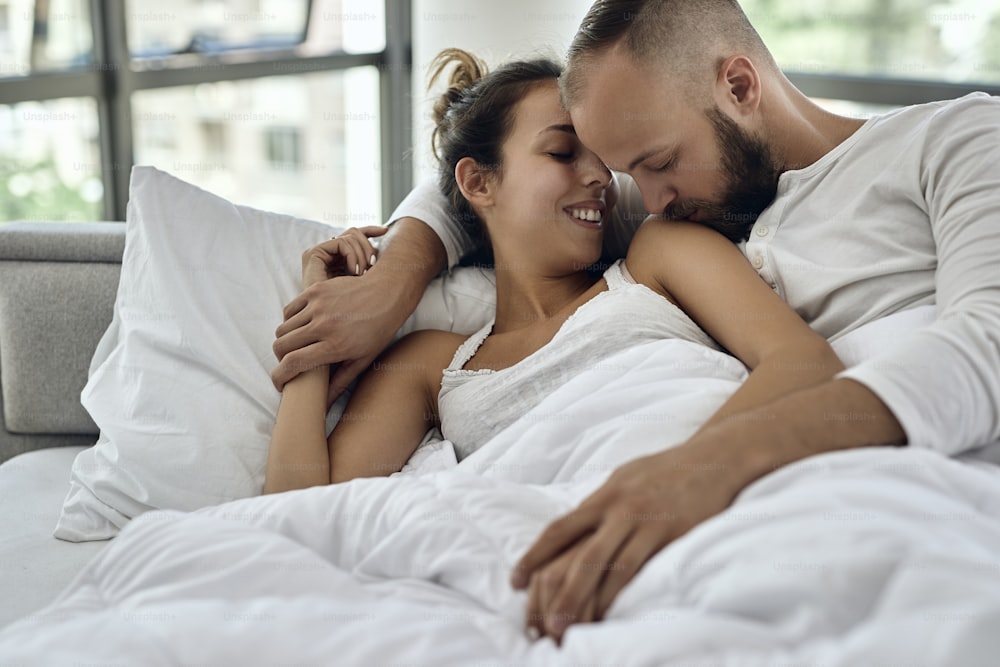 Young couple in love lying embraced on a bed. Man is kissing his girlfriend on the shoulder.