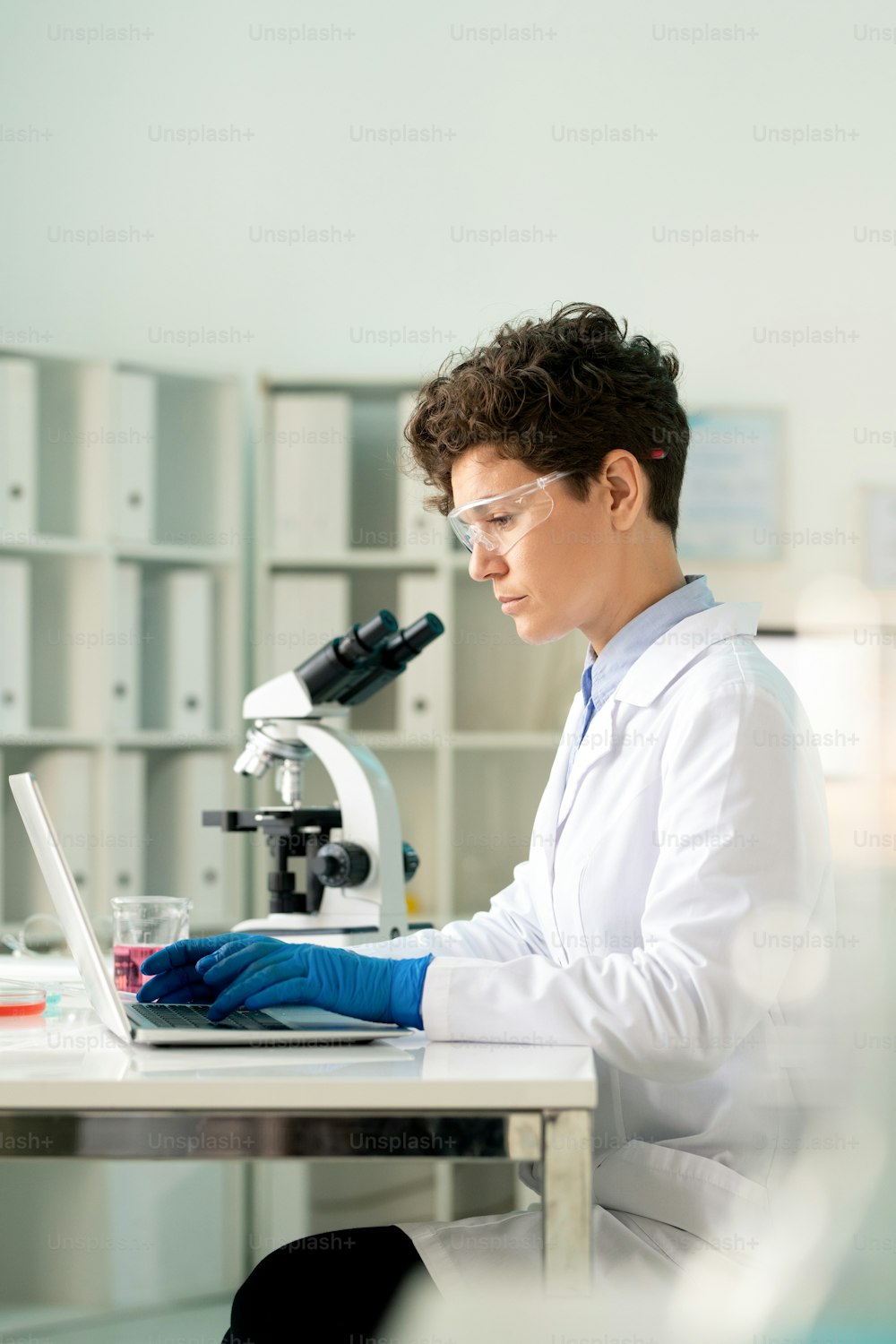 Konzentrierter Virologe mit Schutzbrille und Handschuhen tippt auf dem Laptop während der Arbeit an der Coronavirus-Forschung im Labor