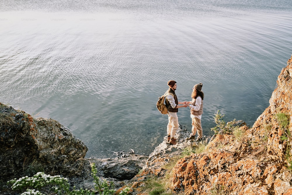 Young affectionate romantic couple holding by hands while standing on stone rock by waterside and looking at one another