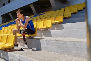 Retrato de un atleta adaptado con pie protésico sentado solo en el estadio vacío en un día soleado de verano, sintiéndose deprimido. Cuerpos Reales, Perseverancia, Coraje, Confianza, Poder, Concepto de Apoyo